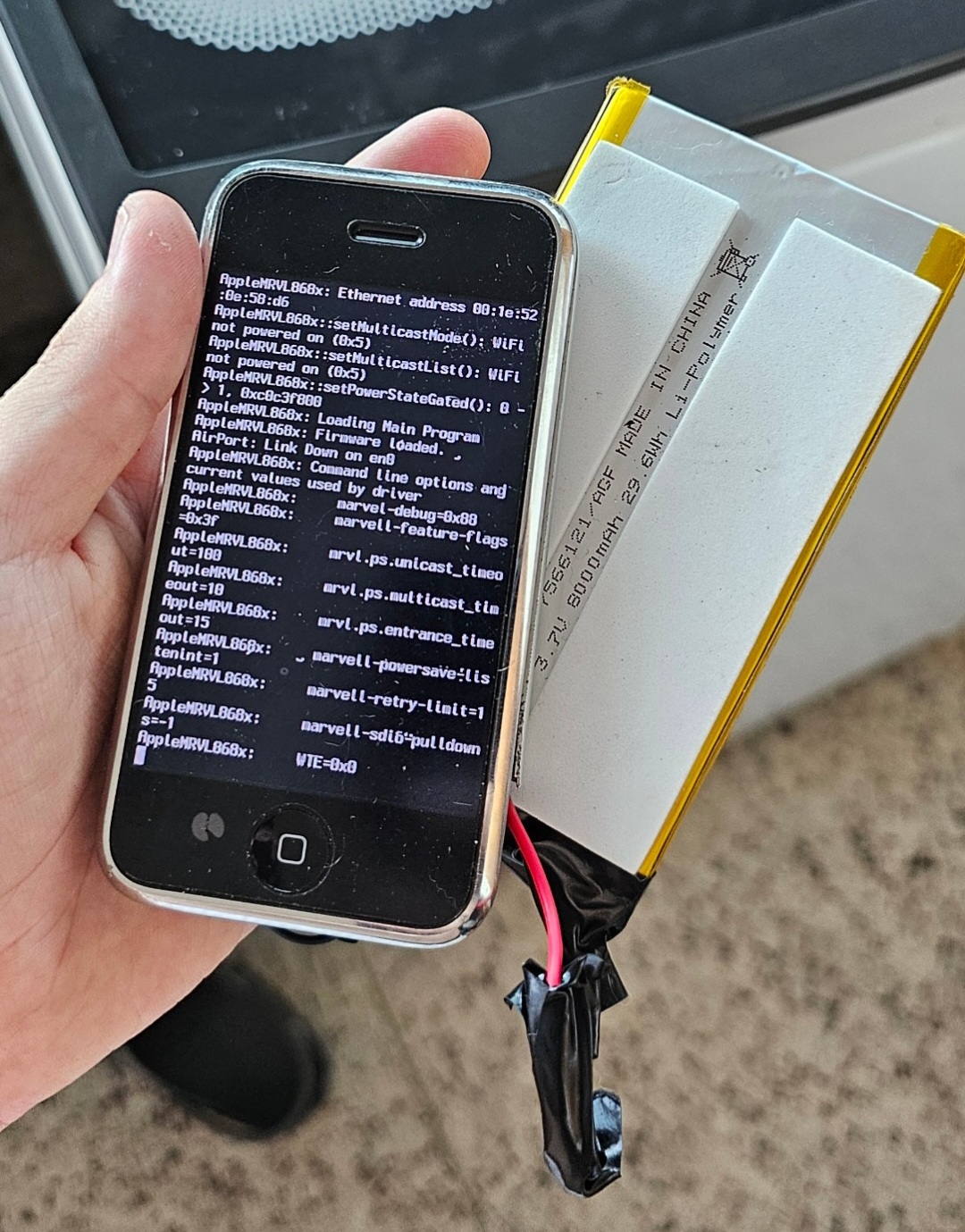 iPhone 2G booted showing AirPort debug console output next to new battery